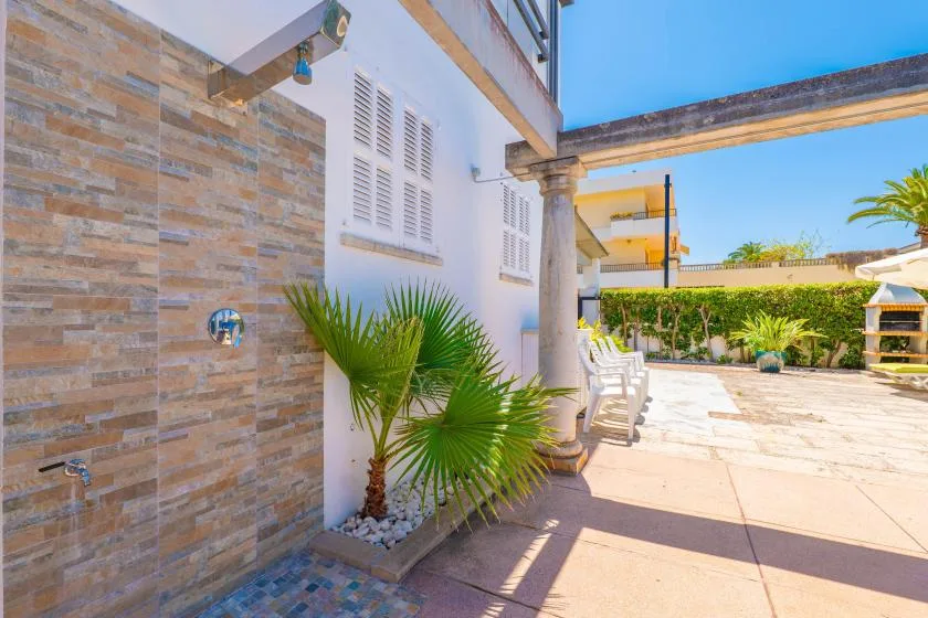 Un patio exterior con una pared de ducha revestida de azulejos, asientos blancos y una palmera en maceta.