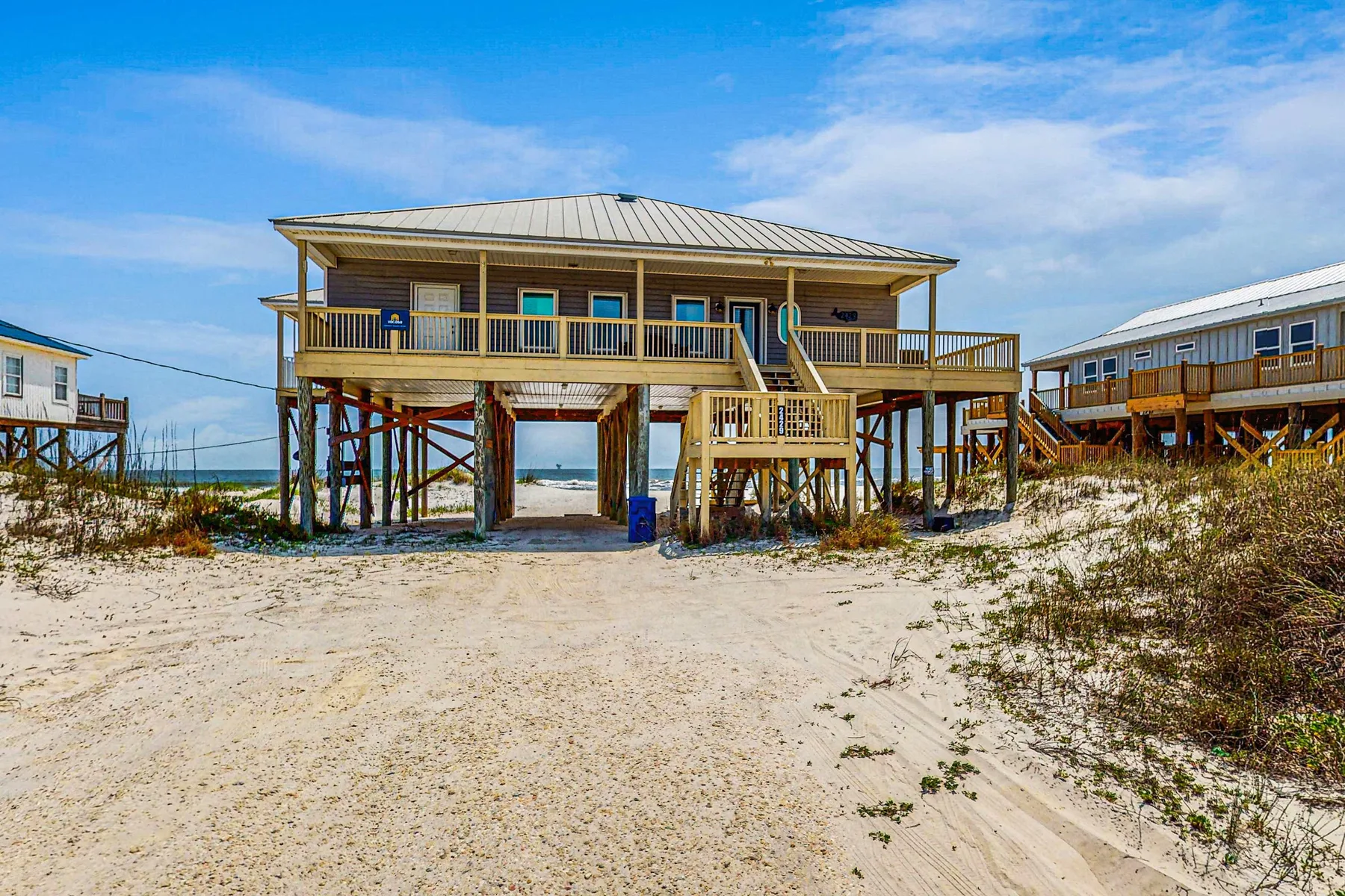 Una casa de playa elevada se alza sobre la arena bajo un cielo azul.