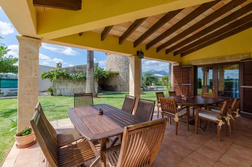 Una zona cubierta de comedor al aire libre con mesas y sillas de madera da a un jardín.