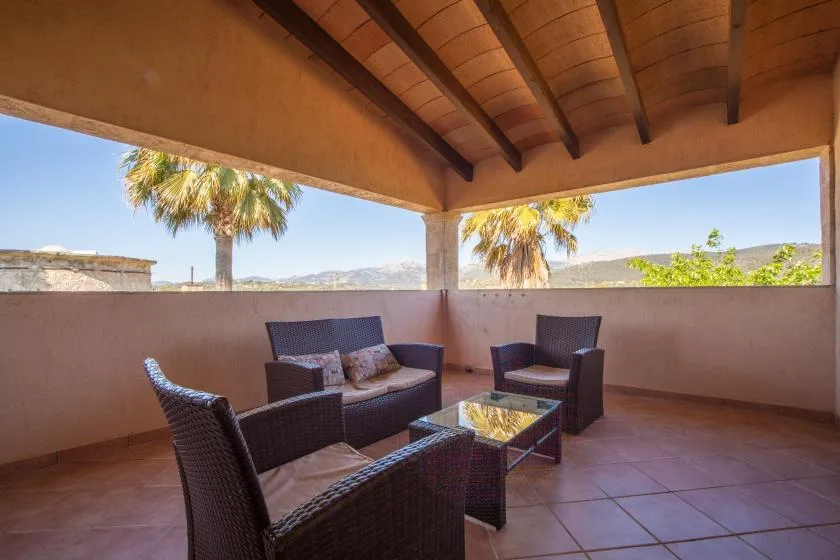 Un patio cubierto con asientos alrededor de una mesa baja da a palmeras y colinas lejanas.