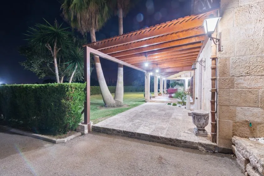 Un pasillo exterior cubierto con vigas de madera, suelo de piedra y vegetación por la noche.