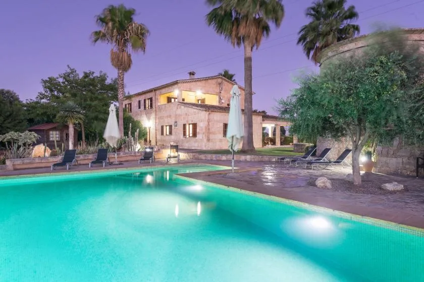 Una casa de piedra iluminada da a una piscina turquesa rodeada de palmeras.