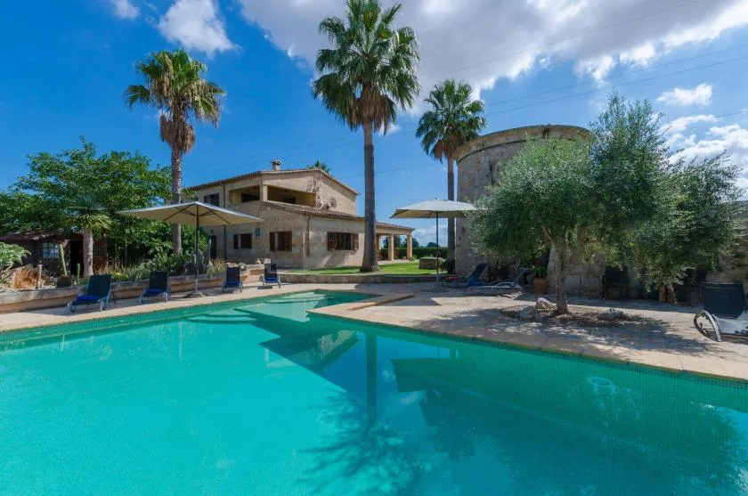 Una piscina al aire libre junto a un edificio de piedra, a la sombra de palmeras.