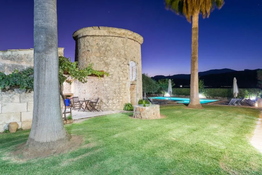 Una zona ajardinada junto a la piscina, con una torre de piedra, palmeras y asientos al aire libre.