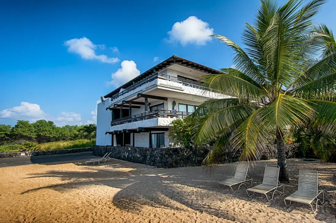 A beachfront building with balconies stands beside sandy shore and palm trees.