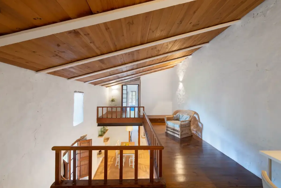 Un luminoso descansillo tipo loft con barandilla de madera, vigas vistas y una silla de mimbre.