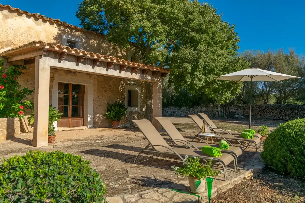 Un patio de piedra junto a una casa tiene asientos reclinables y una sombrilla grande.
