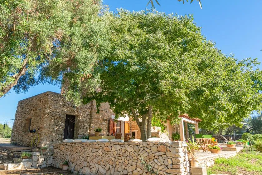 Una casa de piedra está bajo árboles frondosos, con un muro bajo de piedra al frente.