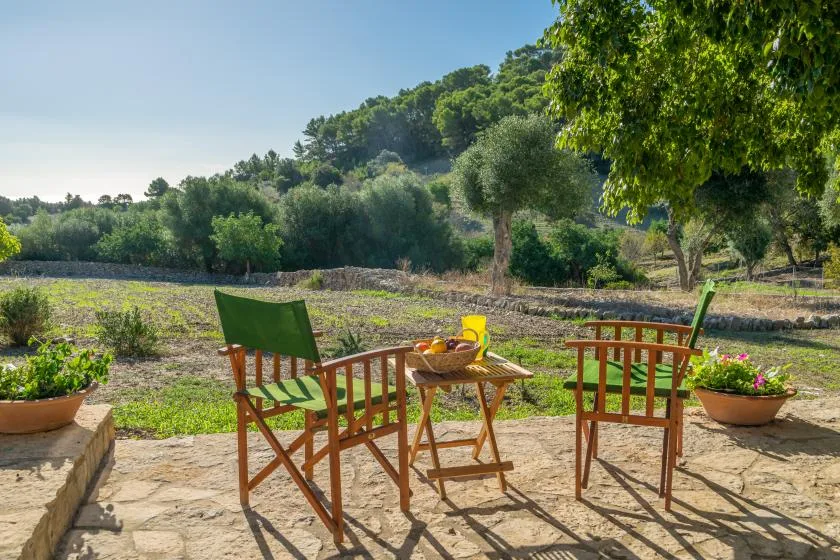 Una zona de estar al aire libre con sillas de madera y una mesa pequeña da a un paisaje verde.
