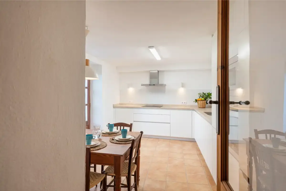 Una cocina y comedor luminosos con gabinetes blancos y una mesa de madera.