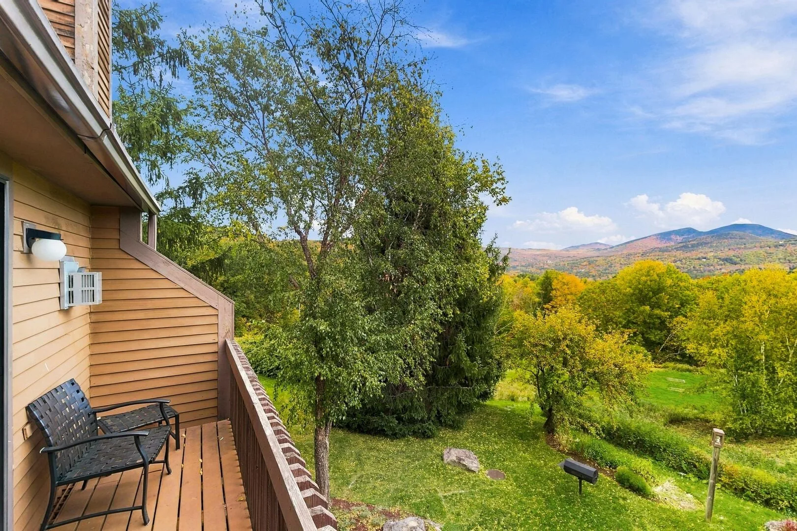 A wooden balcony overlooks a green yard, leafy trees, and distant hills.