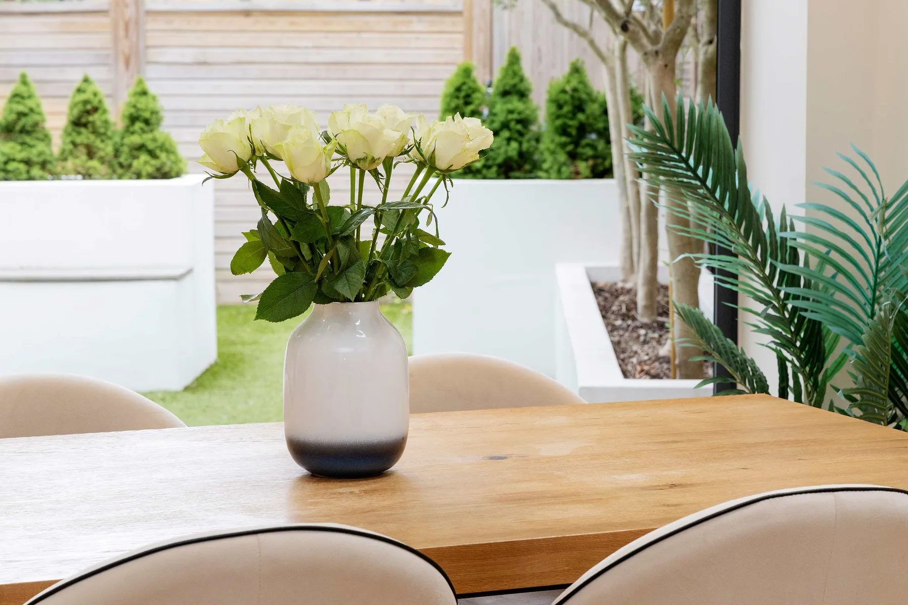 A wooden dining table with a vase of flowers sits beside a planted courtyard.