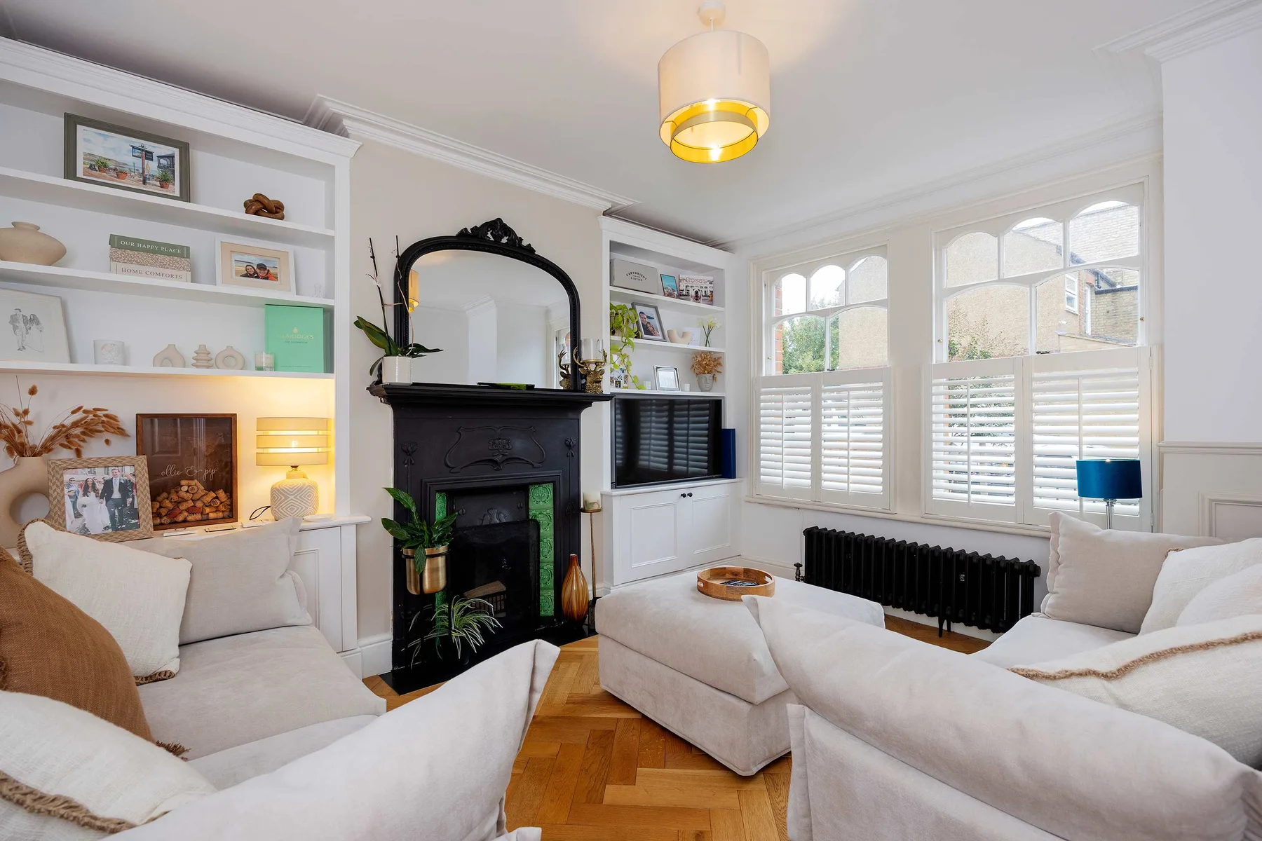 Una luminosa sala de estar con asientos blancos, estantes empotrados y grandes ventanas con contraventanas.