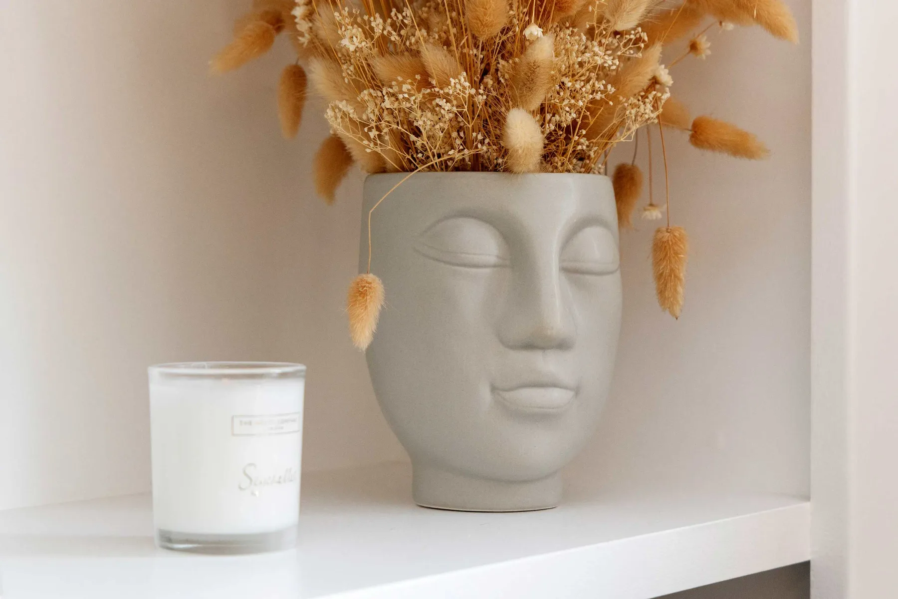 Un jarrón decorativo con forma de rostro y flores secas está sobre una repisa blanca junto a una vela.