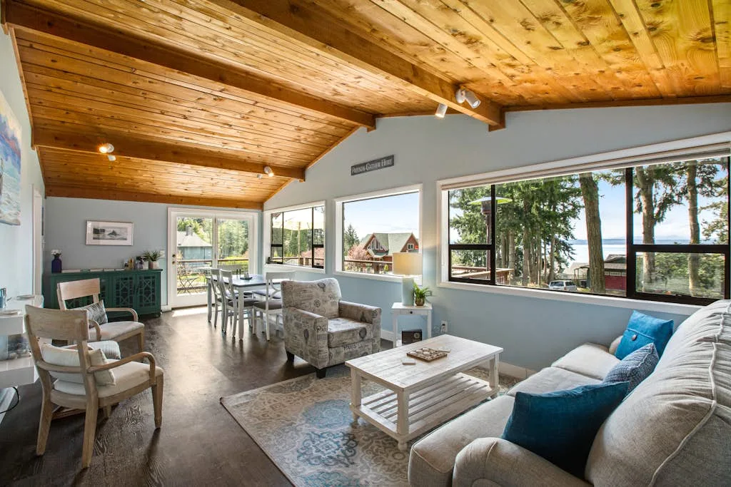 A bright living and dining area has wood ceilings and large windows overlooking trees.