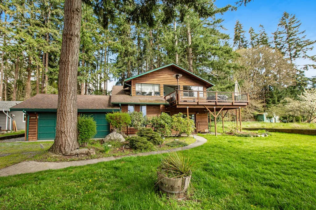 Una casa de madera está entre árboles altos, junto a un césped verde y una terraza elevada.