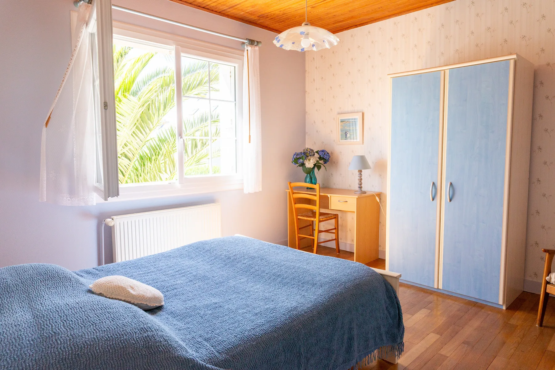 A bedroom with a blue bedspread, a wardrobe, and a bright window.