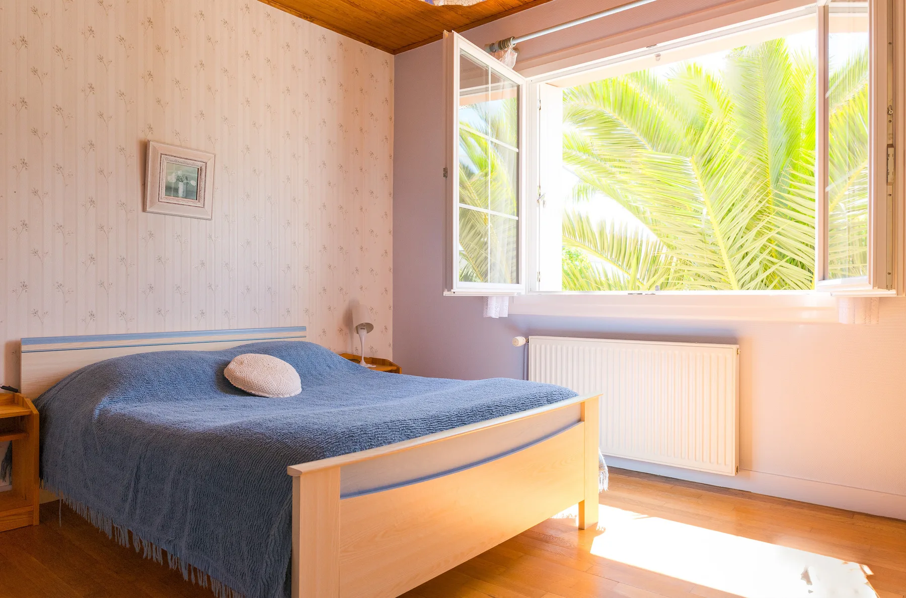 A bedroom with a bed beside an open window overlooking palm leaves.