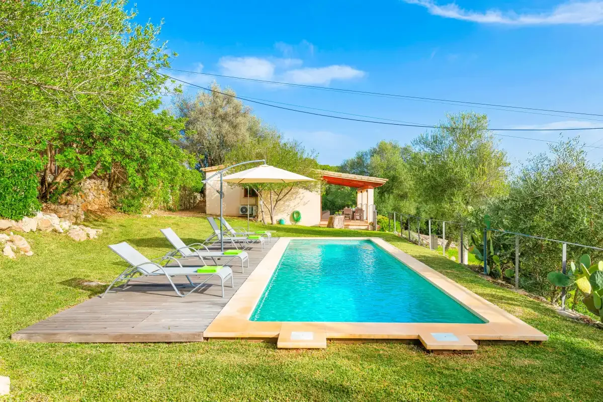 A swimming pool sits beside a wooden deck with sun loungers in a green garden.