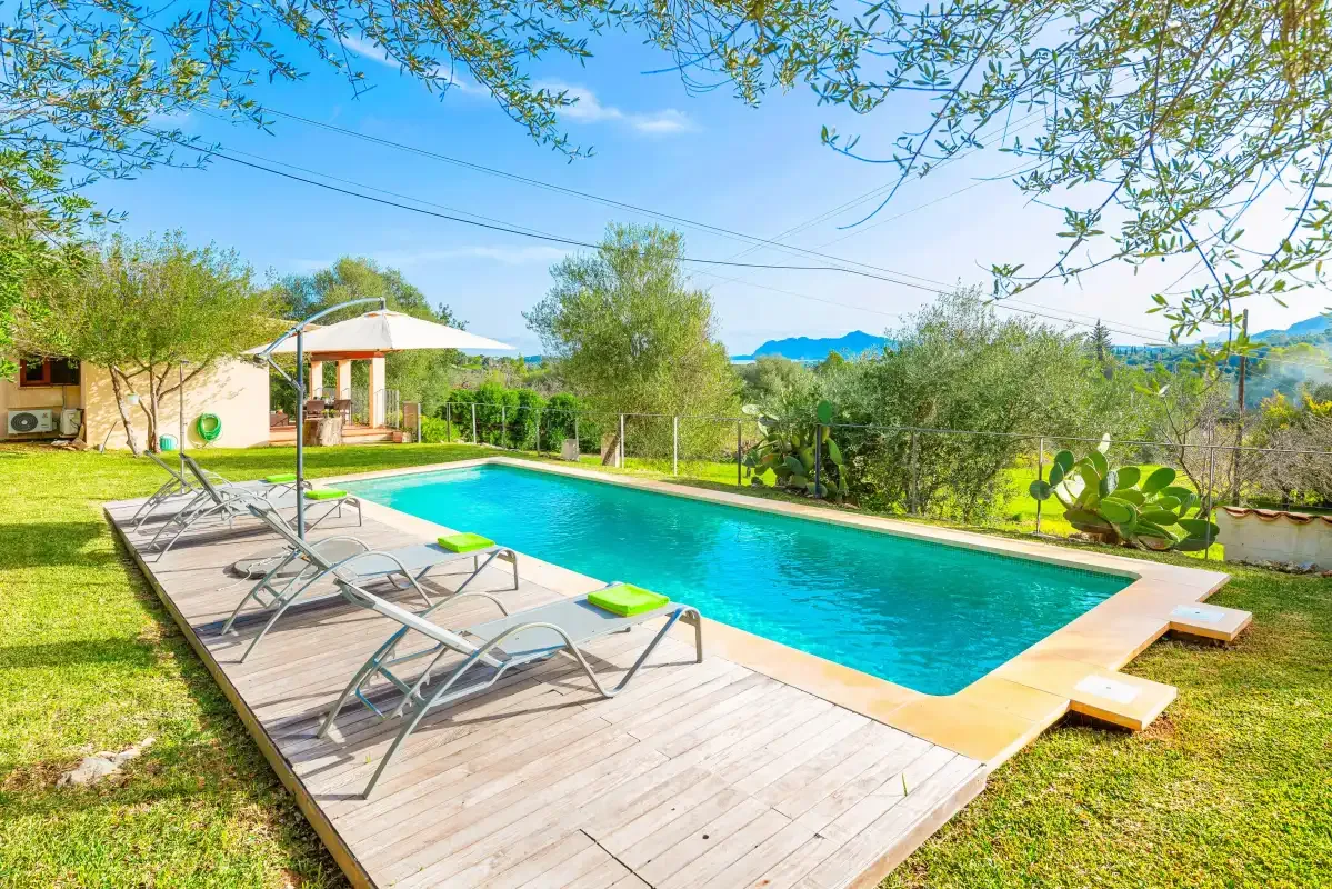 An outdoor swimming pool beside a wooden deck with reclining seating and surrounding greenery.