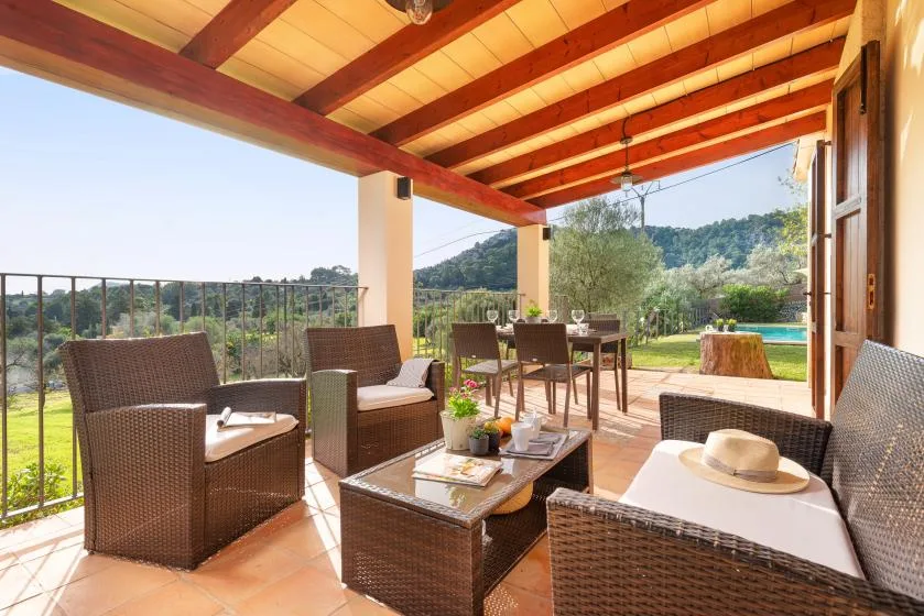 A covered patio with wicker seating and a dining table overlooks a green landscape.