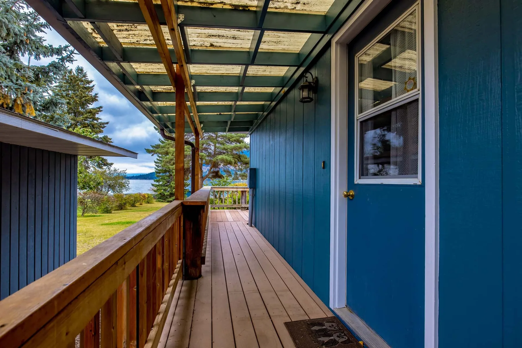 A covered wooden porch runs alongside a blue building, with a grassy yard beyond.