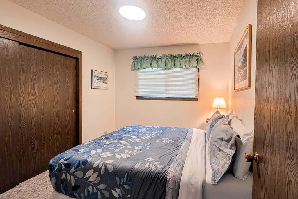 A bedroom with a bed beneath a window and a lit bedside lamp.