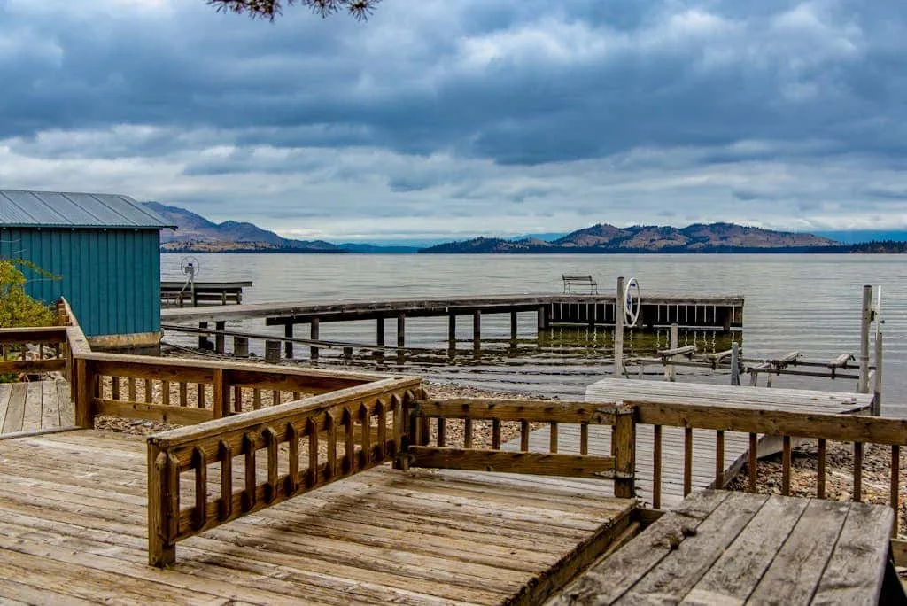 A wooden lakeside dock with railings extends over the water beneath a cloudy sky.