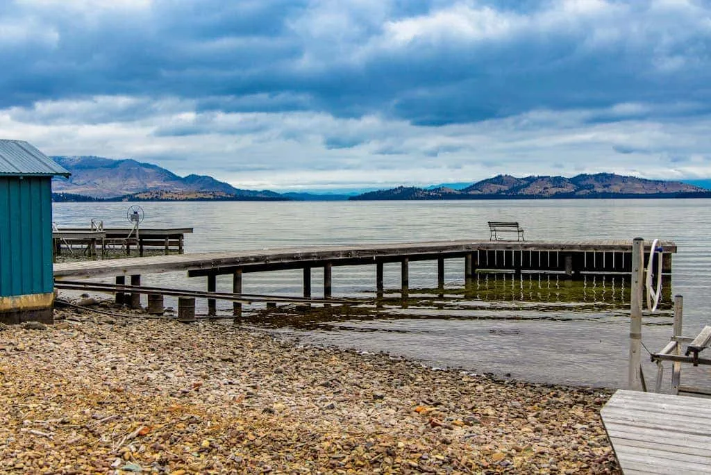 A lakeside pier extends over calm water beside a pebbled shore.