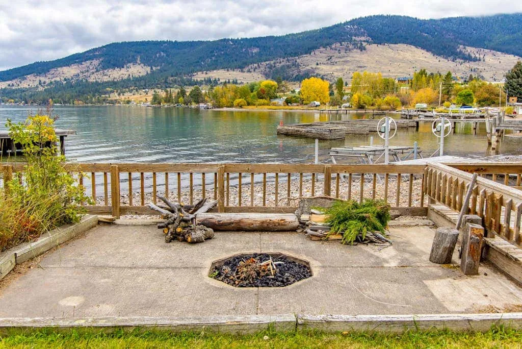 A lakeside patio overlooks the water, with a fire pit in the foreground.