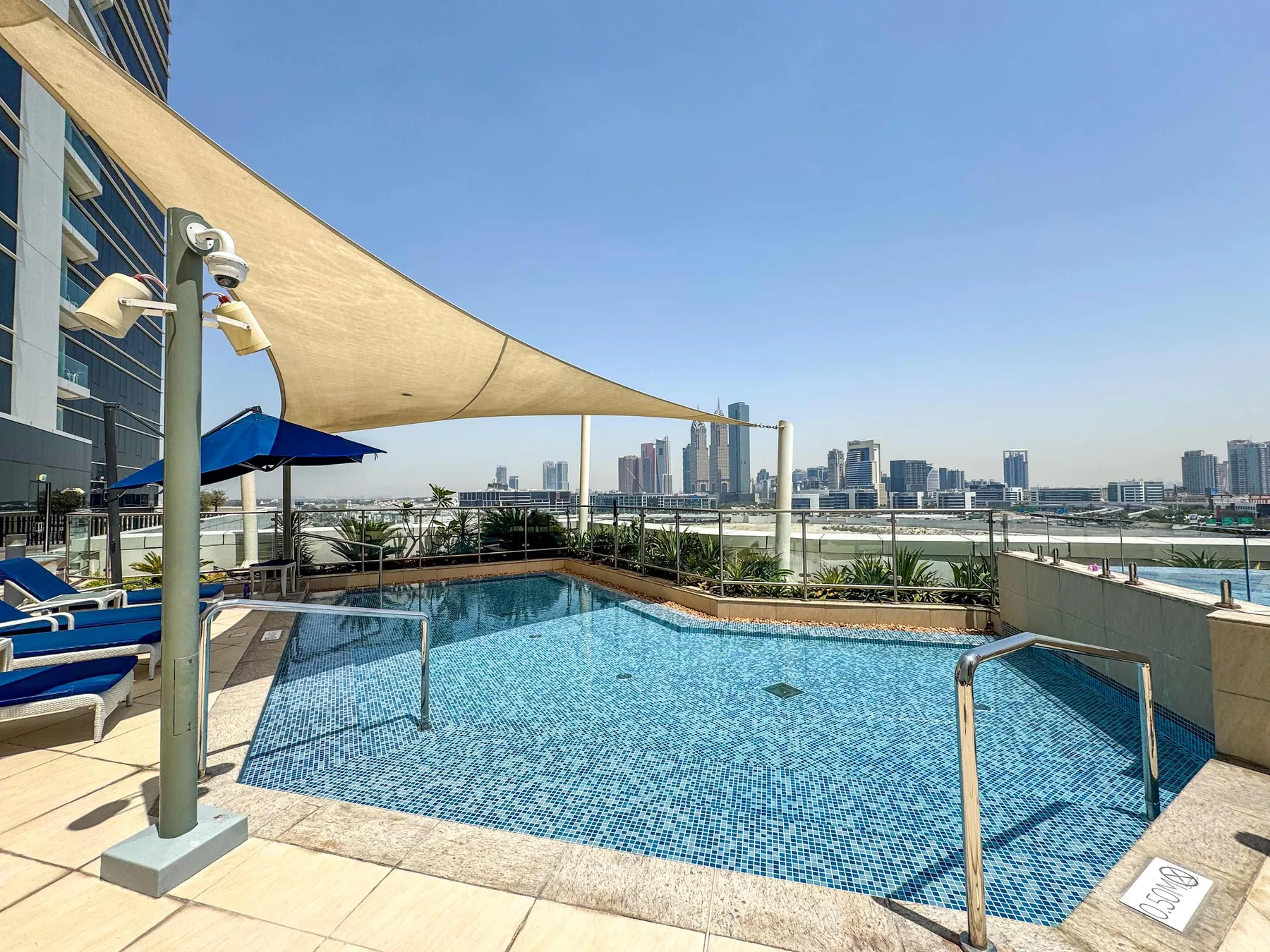 A rooftop swimming pool sits beside a shaded deck with a city skyline in the distance.