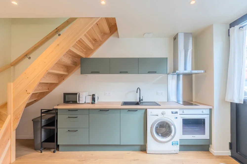 A compact kitchen beneath a wooden staircase, with green cabinets and appliances.