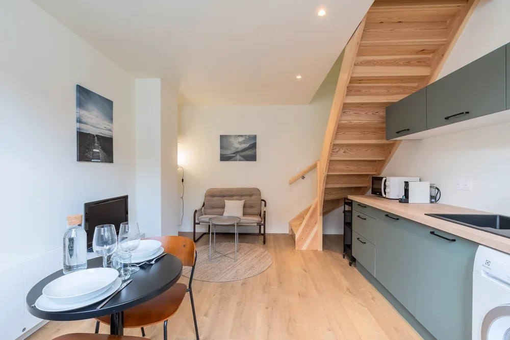 A bright open-plan room with a dining table, kitchenette, and wooden staircase.