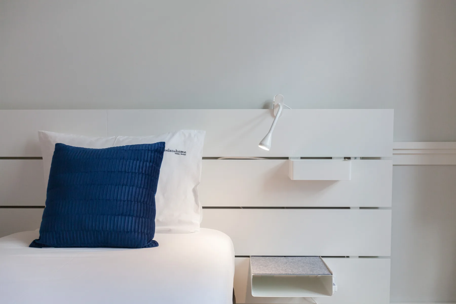 Un dormitorio con una cama blanca, un cojín azul y una lámpara de lectura en la pared.