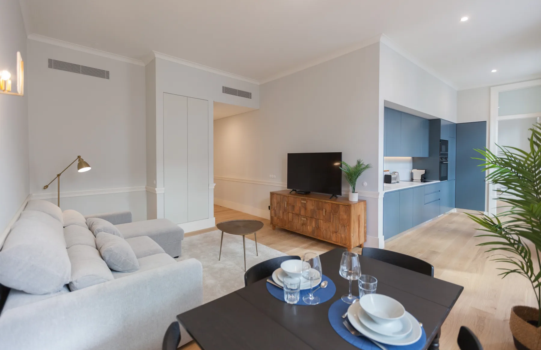 Sala de estar y comedor con asientos claros, televisor y cocina azul.