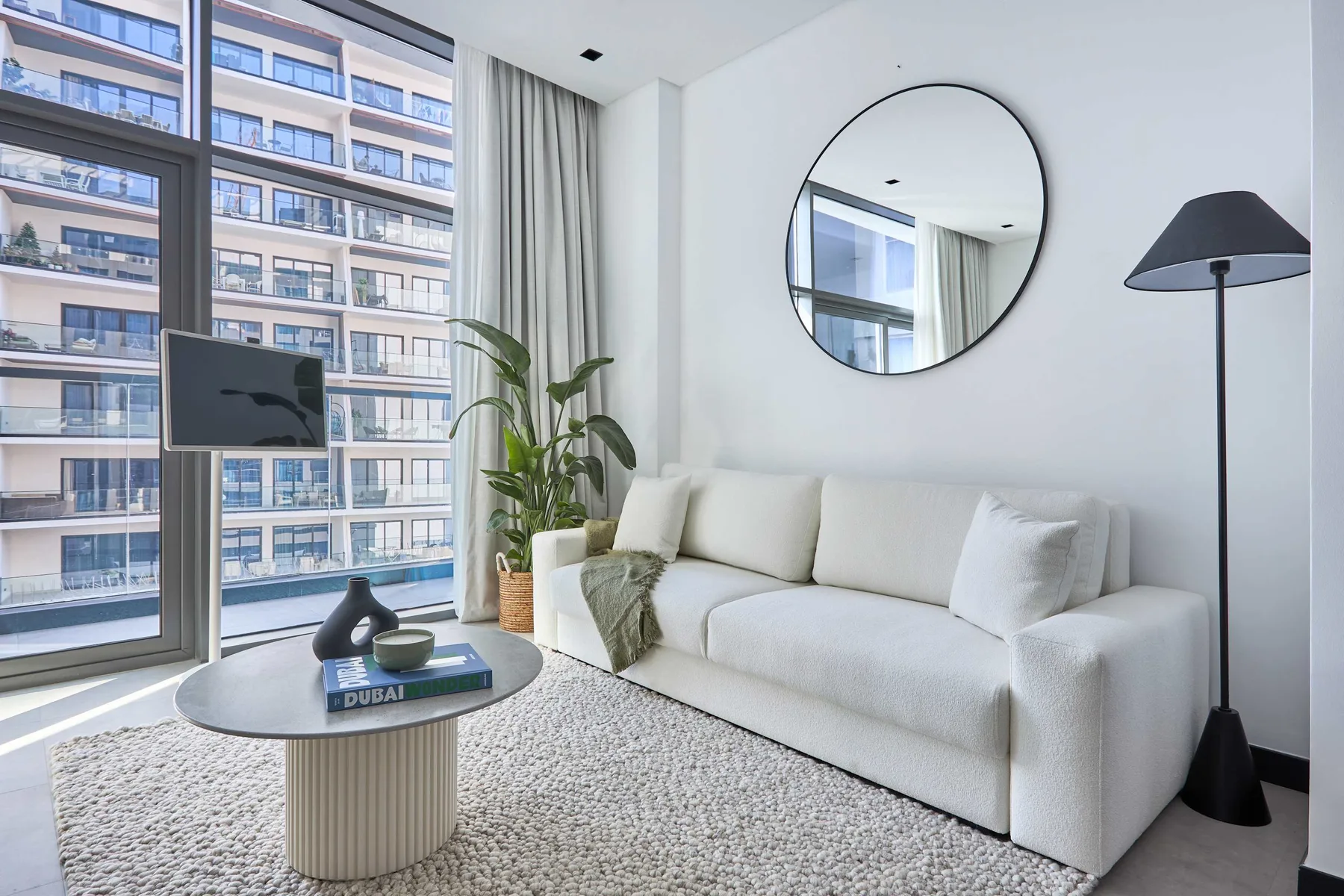 A bright living room with white seating, a round mirror, and large windows.