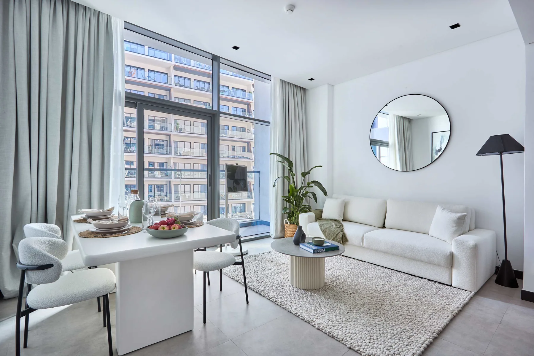 A bright living room with white seating, a dining table, and large windows.