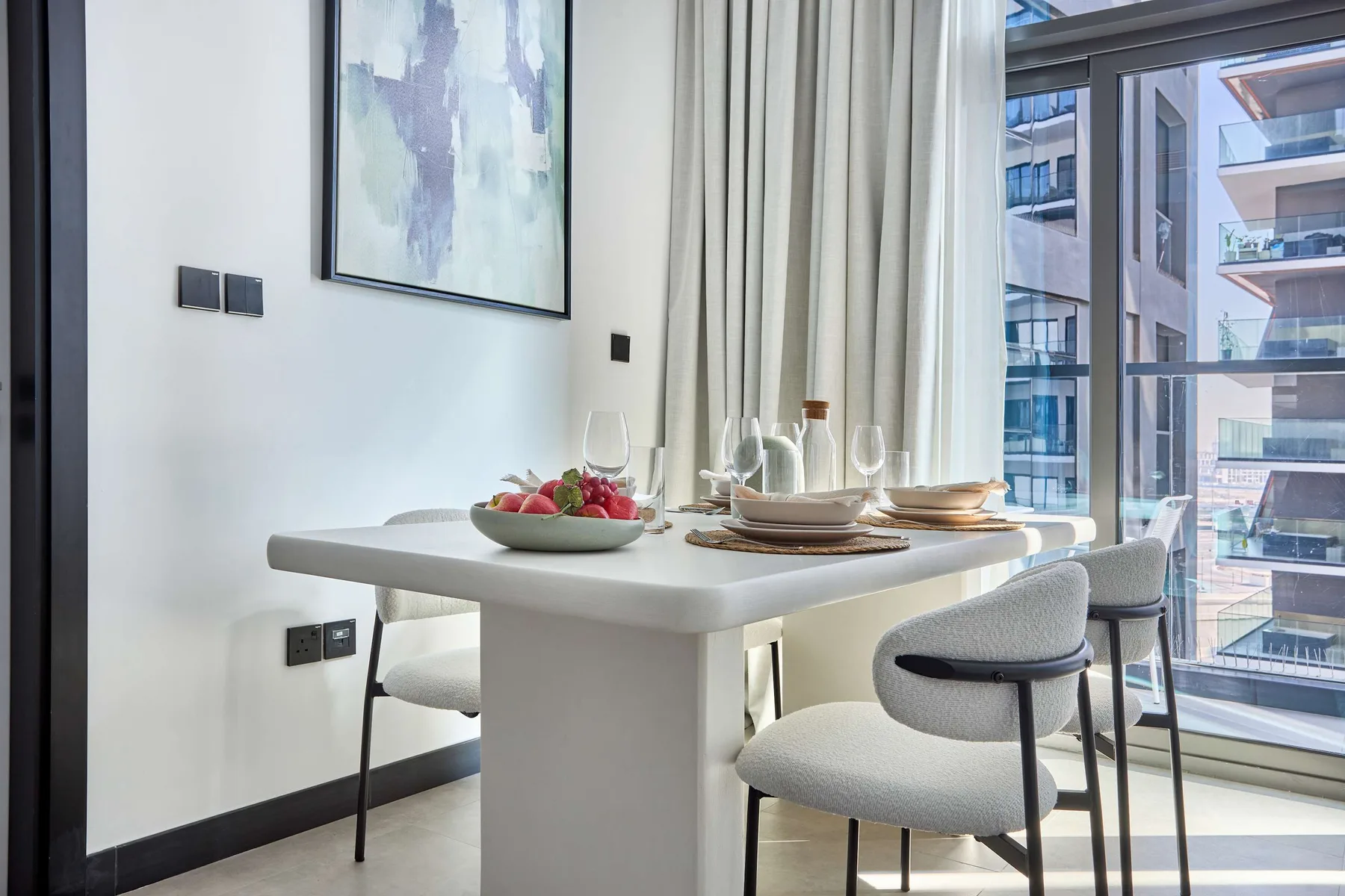Comedor con la mesa puesta junto a grandes ventanas con vistas a edificios cercanos.