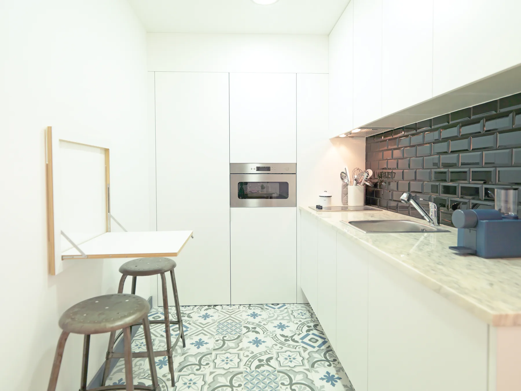 Una cocina moderna y luminosa con una encimera larga, azulejos oscuros y suelo estampado.