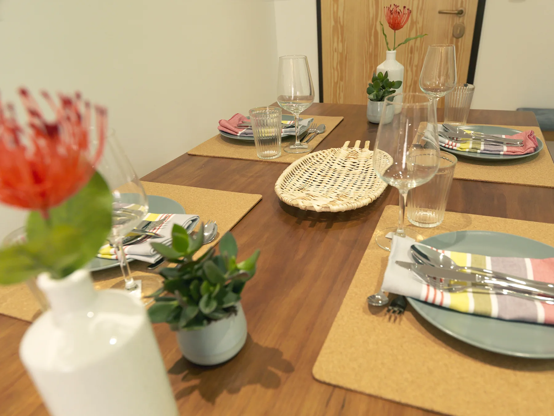 Una mesa de comedor está puesta con manteles individuales, platos, vasos y pequeñas plantas.