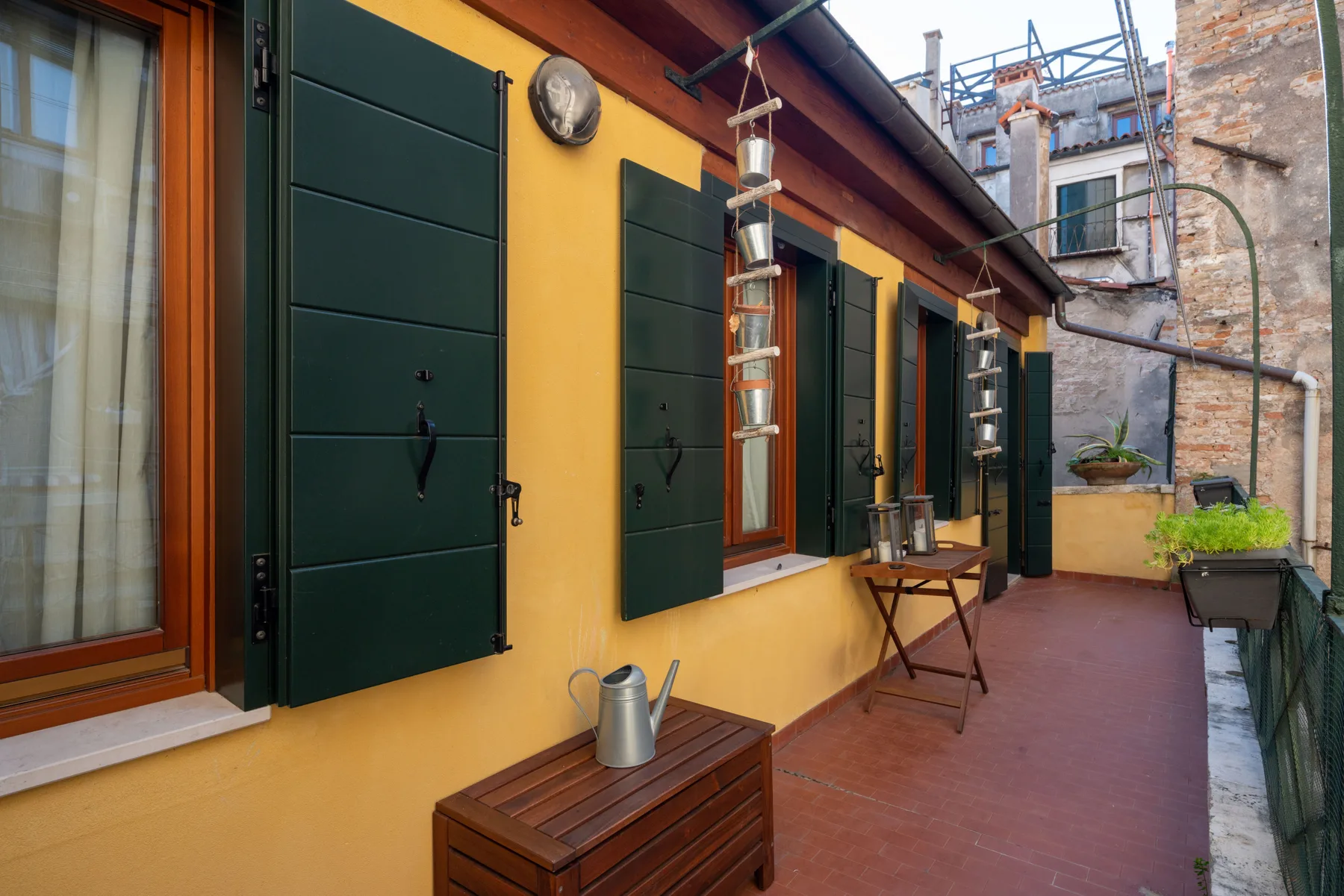Una terraza exterior estrecha bordea una pared amarilla con contraventanas verde oscuro.