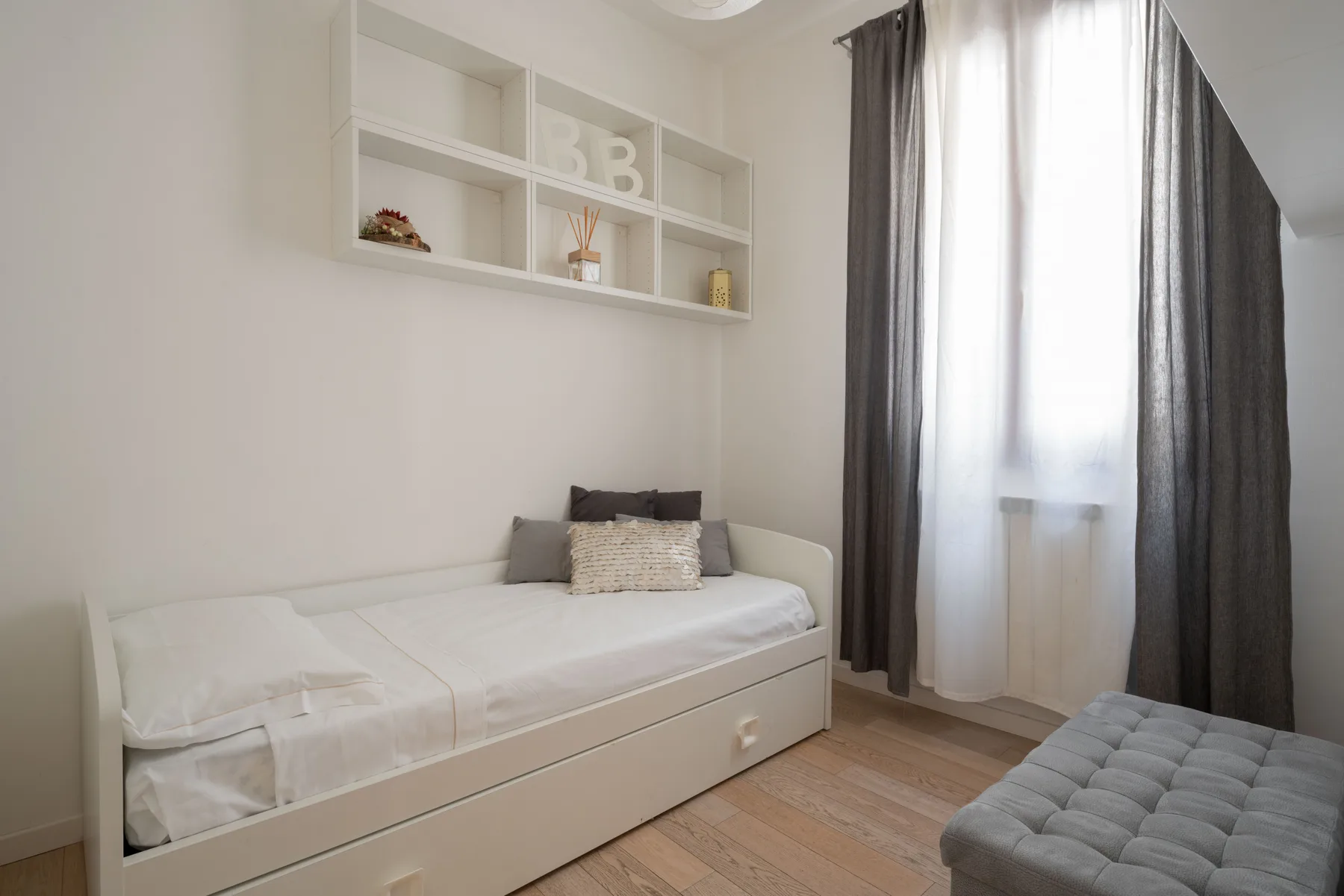 A bright bedroom with a single bed, a high window, and gray curtains.