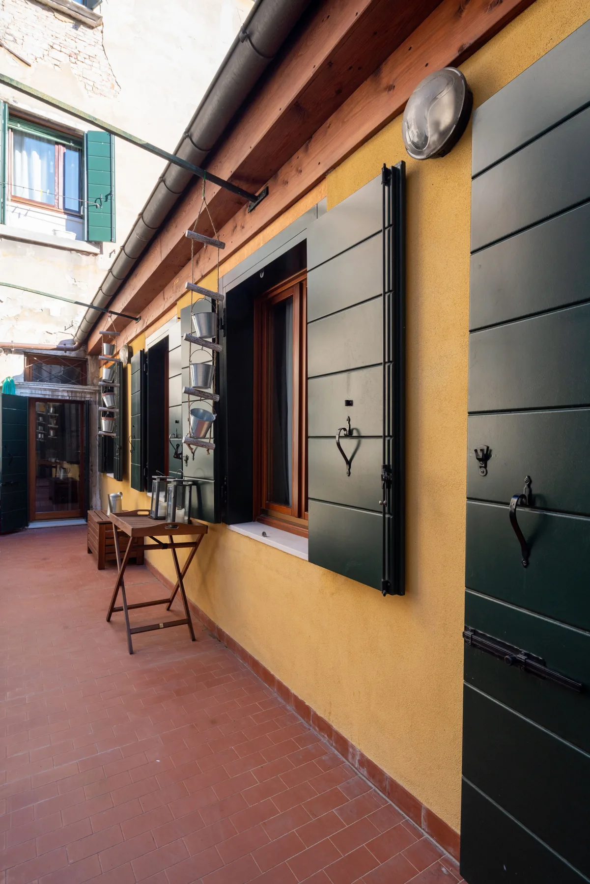 A tiled outdoor passage runs alongside a yellow wall with dark shutters and windows.