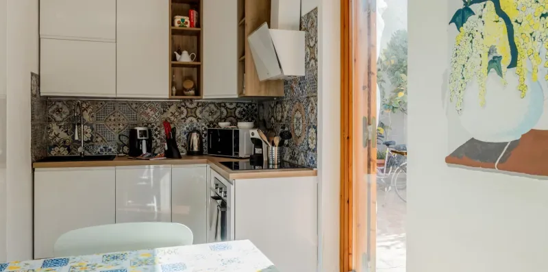 A compact kitchen with pale cabinets, patterned backsplash, and a table in the foreground.