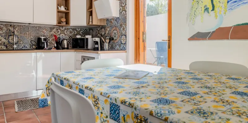 A dining table with a patterned tablecloth sits beside a bright kitchen.
