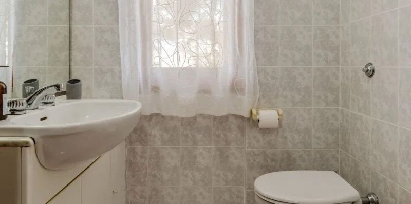 A tiled bathroom with a sink, toilet, and curtained window.