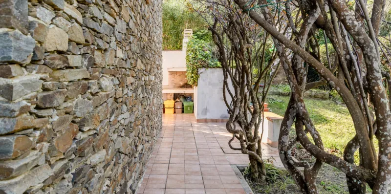 A tiled outdoor walkway runs beside a stone wall and leafy trees.