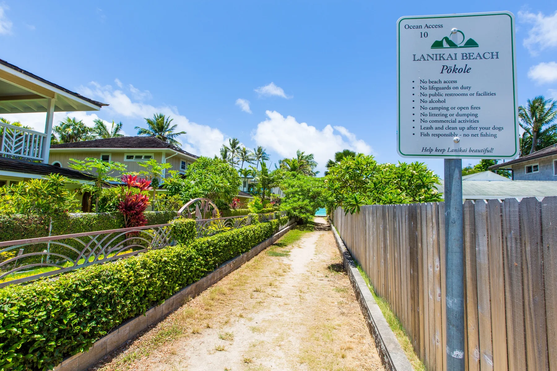 A sandy path runs between tropical gardens and a wooden fence toward the beach.