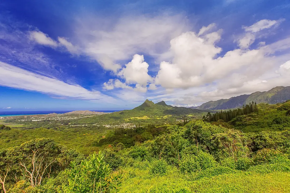 Green hills and dense vegetation stretch toward distant mountains beneath a cloudy blue sky.