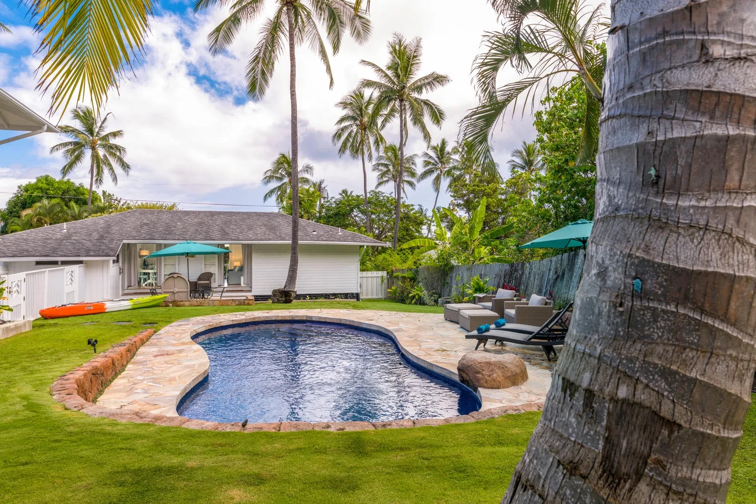 An outdoor pool sits beside a low building, with palm trees and lounge seating nearby.